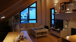 Een gezellige woonkamer met een geruit sofa, een bijzettafeltje en een televisie. Een groot raam biedt uitzicht op het omliggende landschap.