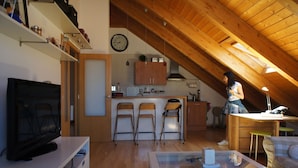 Een gezellige zolderkamer met houten plafond, een televisie, een glazen salontafel en een keukenhoek met stoelen en kastjes.