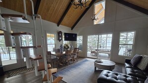 A spacious living area with a wooden ceiling, a black leather sofa, a dining table with chairs, and a large window offering a view of the outdoors. - MT dreams create memories that last a lifetime ❄️ Winter Land ☀️ Summer serenity (Marion)
