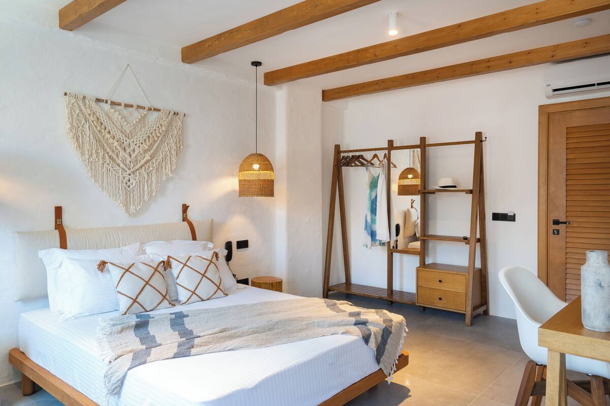A bedroom with a bed, bedside tables, a hanging macrame wall decoration, a wooden shelf with clothes, and a white chair.