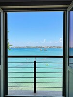 Vista desde un balcón con barandilla, sobre un mar azul y una costa lejana con edificios.