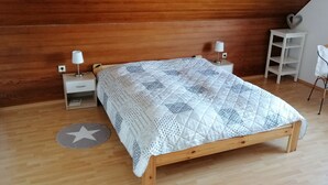 Un dormitorio con una cama de madera, dos mesitas de noche con lámparas, una silla blanca y un estante pequeño blanco.