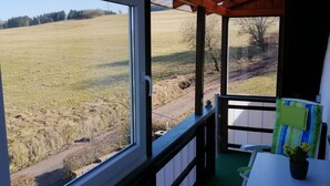 Habitación con un ventanal grande que da a un campo cubierto de hierba. Hay una mesita con una planta y una silla con cojín a rayas.