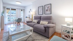 Ett modernt vardagsrum med en grå soffa, ett soffbord i glas och ett vitt sidobord. I rummet finns också en matplats med ett bord och stolar, som syns genom ett stort fönster.