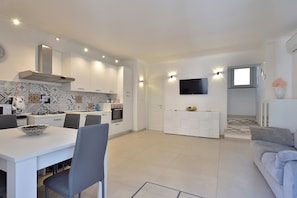 A modern kitchen with white cabinetry, a dining table, and chairs. There is a TV mounted on the wall and a window with a view of the outside. - Sirena Apartment (Minori)