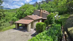 A stone house with a tiled roof, surrounded by lush greenery and a gravel driveway. - Villa Piombona in Anghiari (Anghiari)