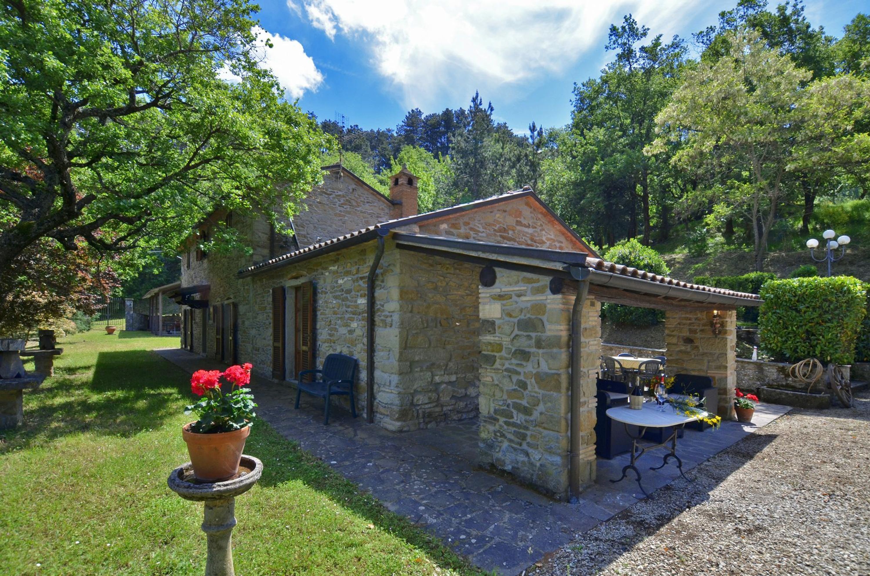 A stone building with a covered patio, surrounded by greenery and trees.