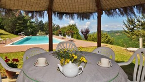 A covered outdoor seating area with a table set for tea, overlooking a swimming pool and lush greenery. - Villa Piombona in Anghiari (Anghiari)