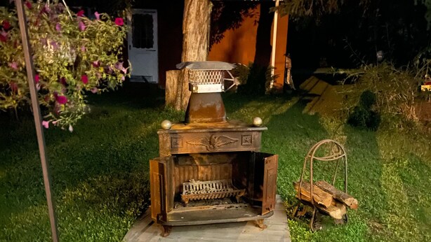 Ein rustikaler Kamin im Freien mit Sitzbank davor, umgeben von Grünpflanzen und einem Baumstumpf.