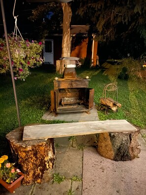 Una chimenea rústica al aire libre con un banco frente a ella, rodeada de follaje y un tocón de árbol.