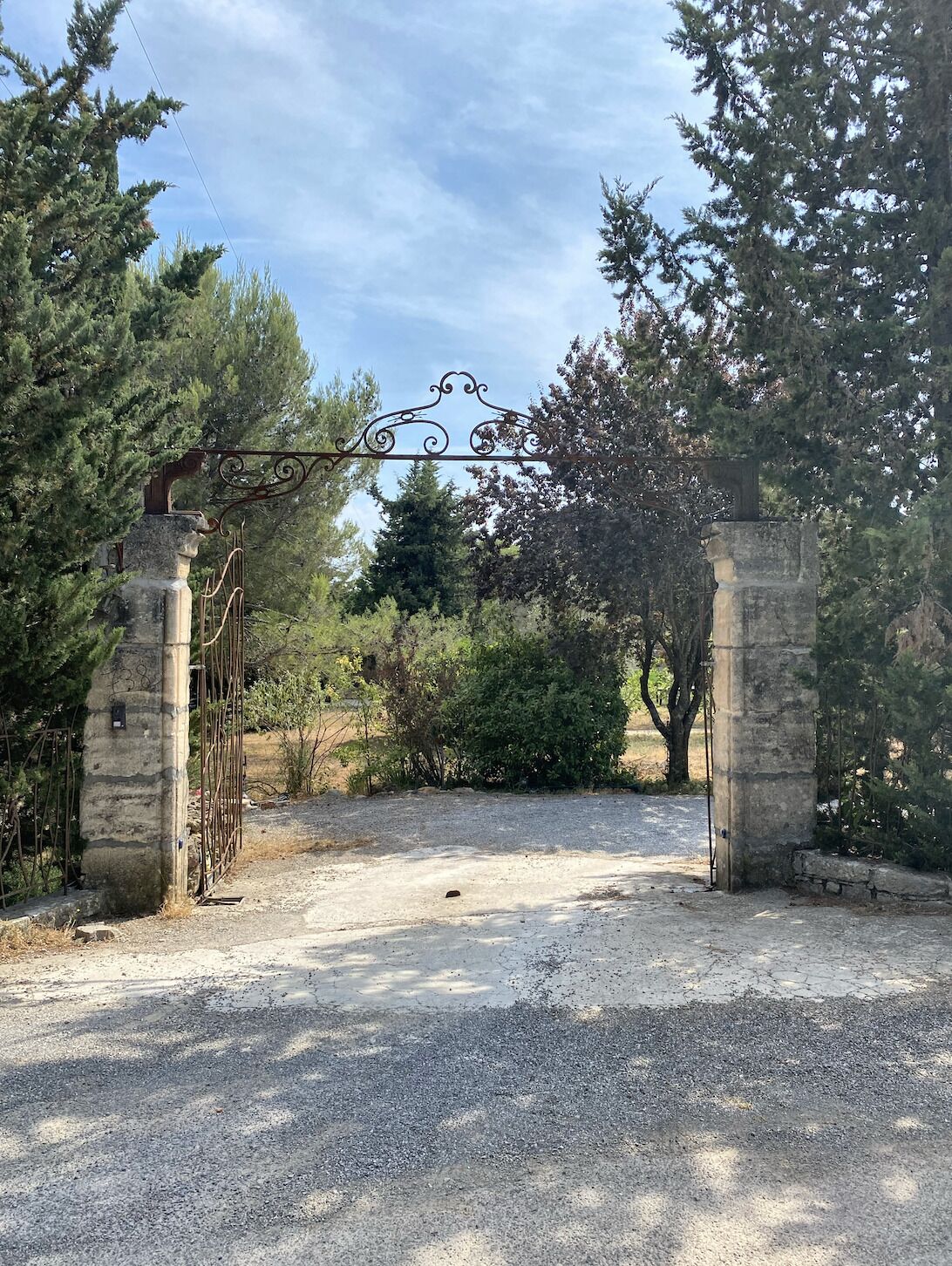 Ein verschlossener Eingang mit einem Metallgitter und steinernen Säulen, umgeben von Bäumen und einem Schotterweg.