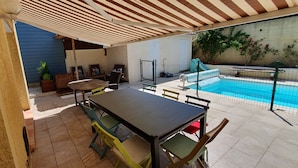 An outdoor patio with a table and chairs, a pool, and a covered seating area.