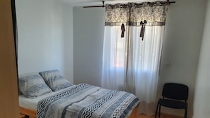A small, single-bed room with a wooden bed frame, a black chair, and a window with sheer curtains.