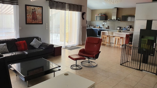 A living room with a black sofa, a red chair, a glass coffee table, and a white side table. There is a kitchen in the background with a dining area.