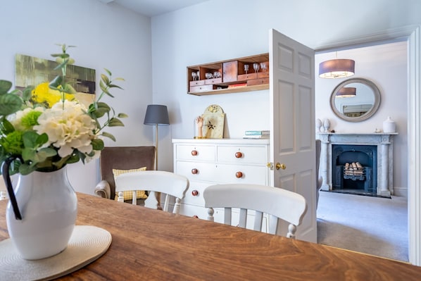 Un coin repas avec une table en bois, des chaises blanches, une commode blanche avec des tiroirs, une horloge et un miroir au mur.