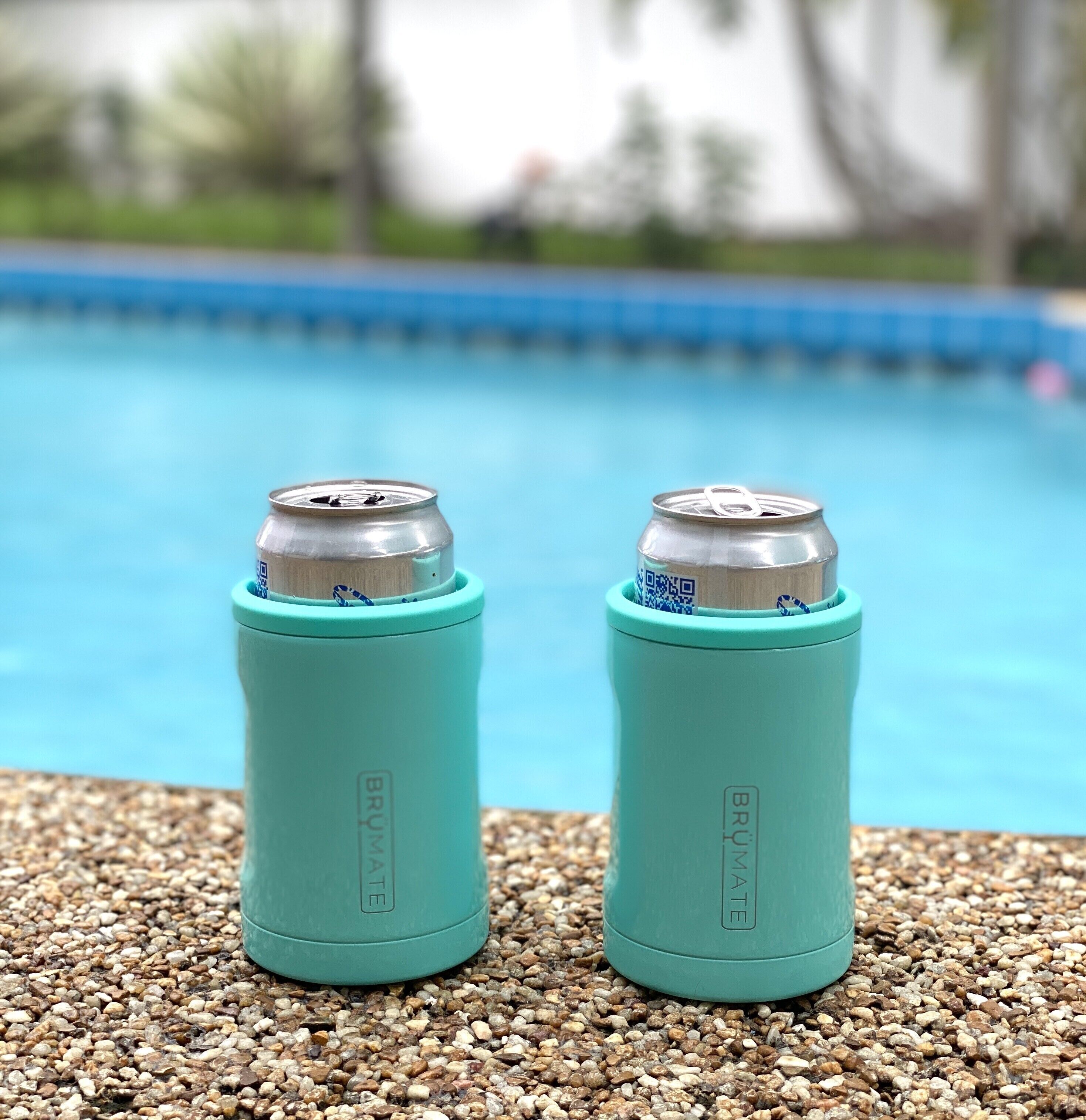 Two teal water bottles with QR codes on a pebbled surface near a pool.