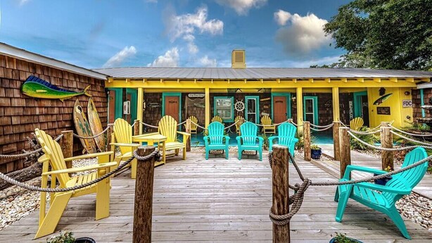 A wooden deck with yellow and turquoise Adirondack chairs, a wooden building with a yellow facade, and a clear blue sky. - Captain's Quarters at Island Breeze of CB (Carolina Beach)