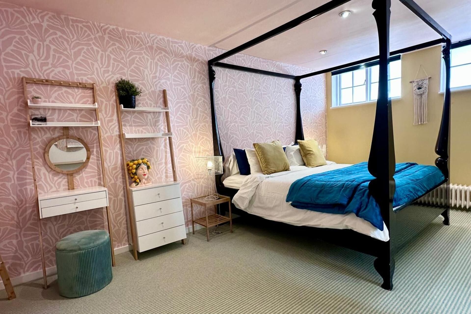 A four-poster bed with a blue and white bedspread, a white dresser with drawers, a round mirror, and a small plant on the dresser.