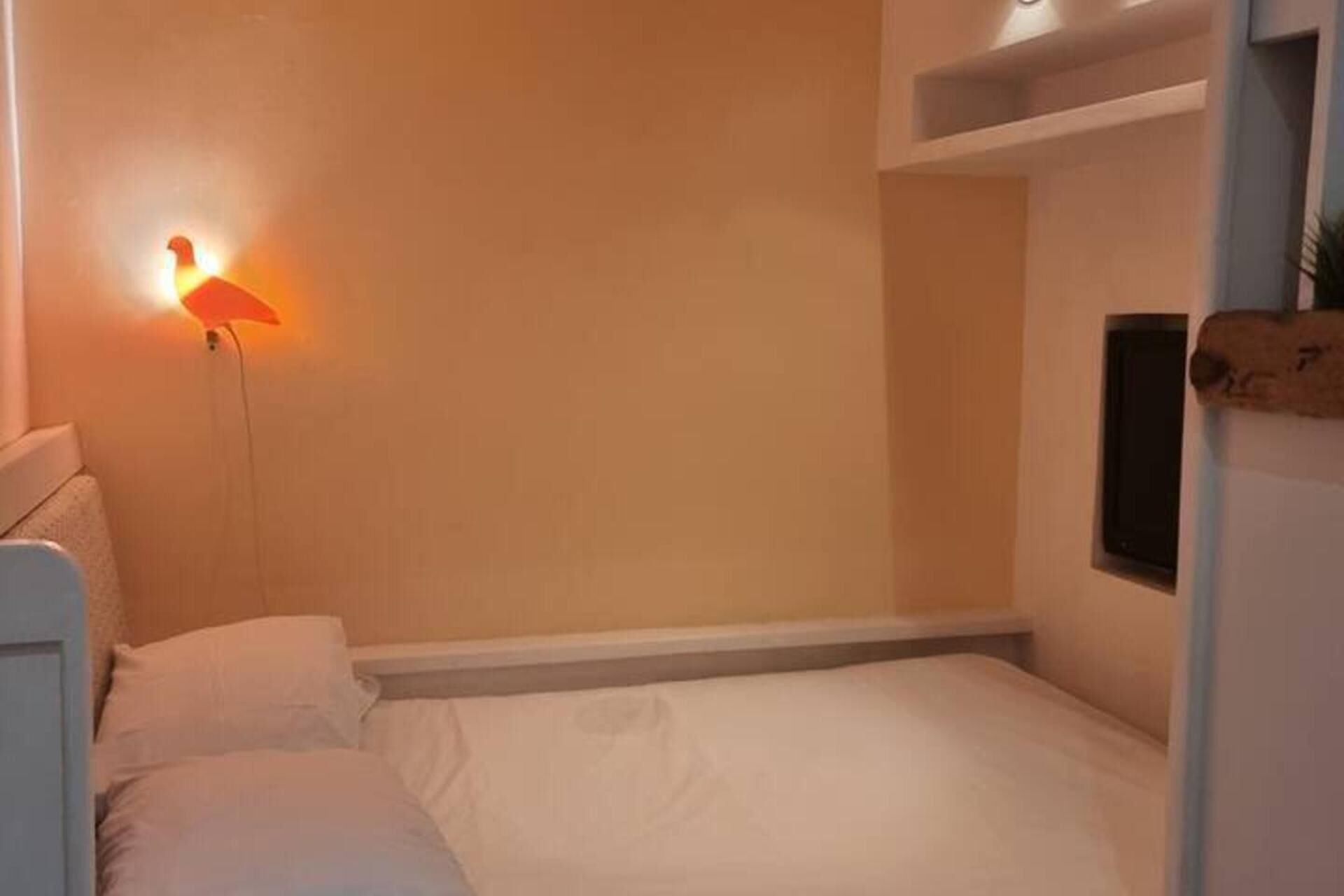 A small, single-bed room with a white bed, a nightstand, and a wall-mounted lamp.