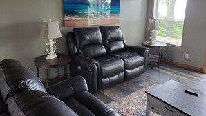 Un salon comprenant un fauteuil inclinable, un canapé, une table basse, une lampe et un tableau représentant une scène de plage.
