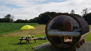 A transparent cylindrical pod with a bed inside, placed on a wooden platform in a grassy field. - Insolite Tonneau Avec Bulle, en Forêt (Pouldergat)