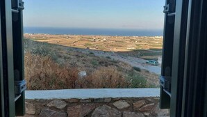 A view through an open window with a stone wall in the foreground, leading to a coastal landscape with fields and a road. - Bluegonia. (Έξω Γωνιά)