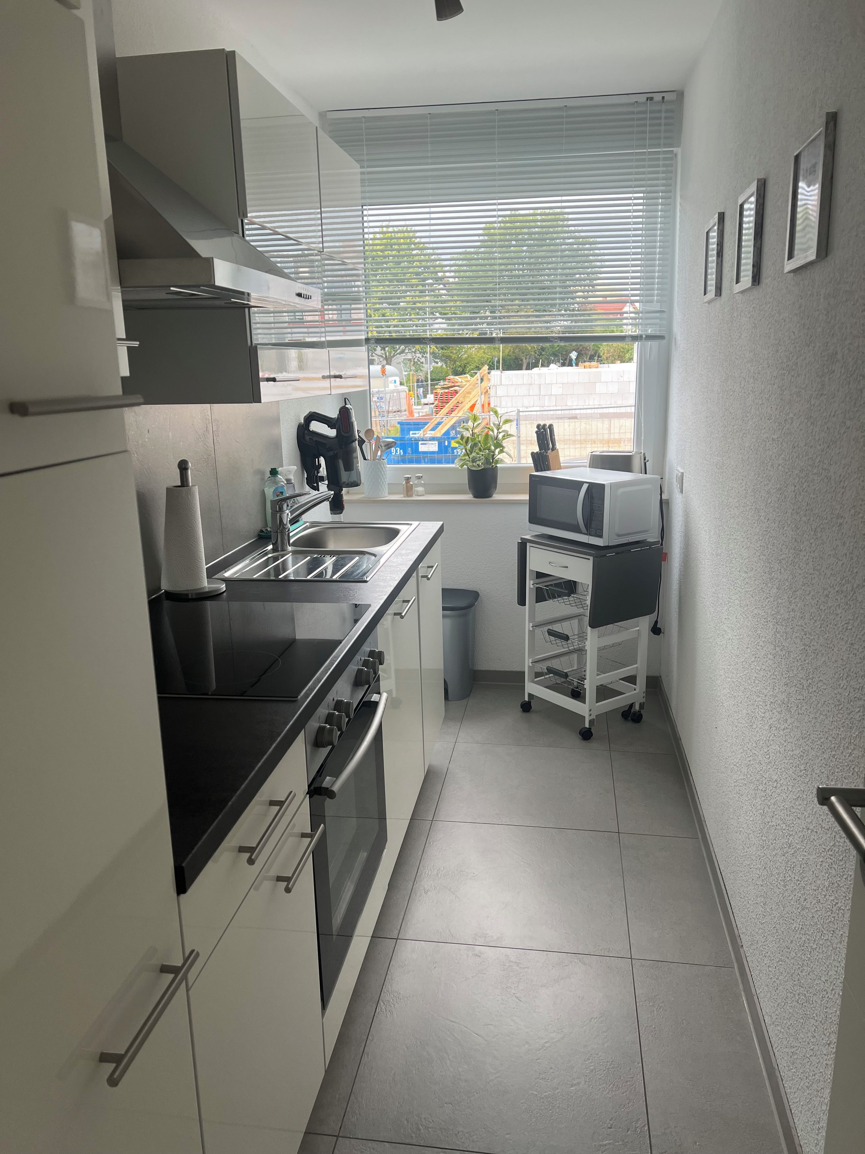 Eine moderne Küche mit Einbauofen, Mikrowelle und Spüle. Ein Fenster mit Jalousien, ein Blumentopf und ein kleiner Wagen mit Küchenutensilien sind ebenfalls vorhanden.