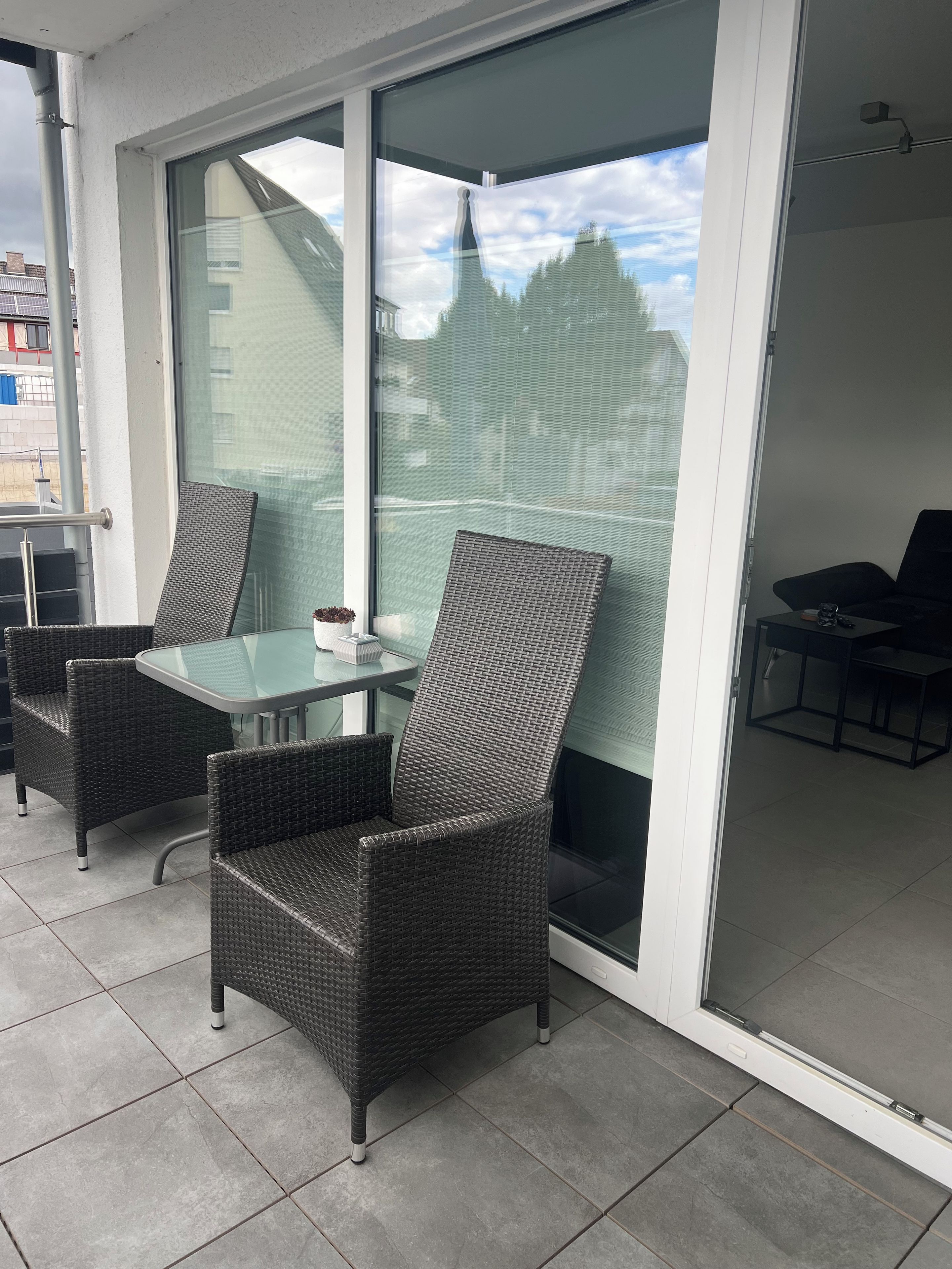 Ein Balkon mit Korbmöbeln, einem Glastisch und Blick auf Gebäude und Bäume.