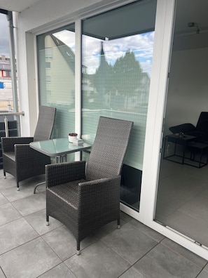 Un balcon agrémenté de meubles en osier, d’une table en verre et offrant une vue sur les immeubles et les arbres.