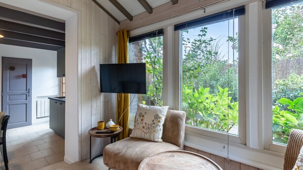 A cozy room with a bed, a TV, a small table, and a window with a view of greenery. - Le Cottage de La Maison Saint Léonard | Lovely Shared Garden in the Heart o (Honfleur)