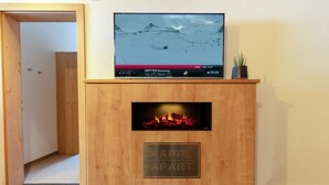 Een houten entertainmentkast met een televisie waarop een besneeuwd berglandschap staat, een open haard en een plaquette met de tekst 'KAPPL APART'.