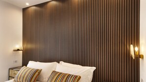 Una habitación de hotel moderna con un panel de pared de madera, una cama bien tendida con sábanas blancas, almohadas a rayas y una mesita de noche con un libro y una lámpara.