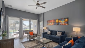 A modern living room with a blue sofa, a coffee table, and a ceiling fan. There is a painting on the wall and a dining area visible through the glass doors.