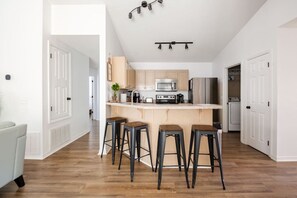 A modern kitchen with a central island, bar stools, and a dining area with a light-colored sofa. - Private (optional) Heated Pool King Beds-NearBeach (Gulf Breeze)