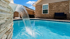 Uma piscina com cachoeira artificial, parede de tijolos e um edifício com janela e banco.