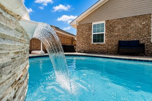 A swimming pool with a waterfall feature, a brick wall, and a building with a window and a bench.
