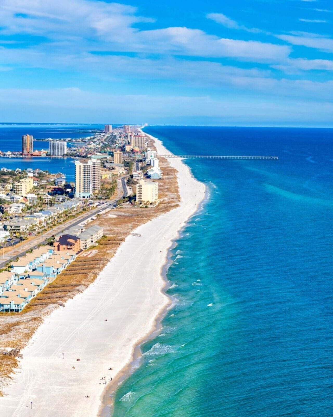 An aerial view of a coastal city with a long sandy beach, a pier extending into the ocean, and a dense cluster of buildings along the shore.