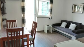 A living room with a sofa, a dining table with chairs, a small white side table, and a window with curtains. - Wohnung für 2 in Einer Villa mit Wunderschönem Meerblick (Karlobag)