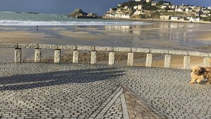 Ein gepflasterter Weg führt zu einem Strand mit Steinbrunnen, mit Blick auf eine Küstenstadt mit Gebäuden auf einem Hügel.