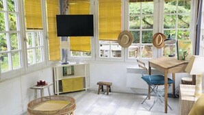 Ein Zimmer mit hoher Decke, großen Fenstern mit gelben Jalousien, einem Fernseher, einem Esstisch mit Stühlen, einem kleinen Hocker und einem Bett mit gemustertem Überwurf.