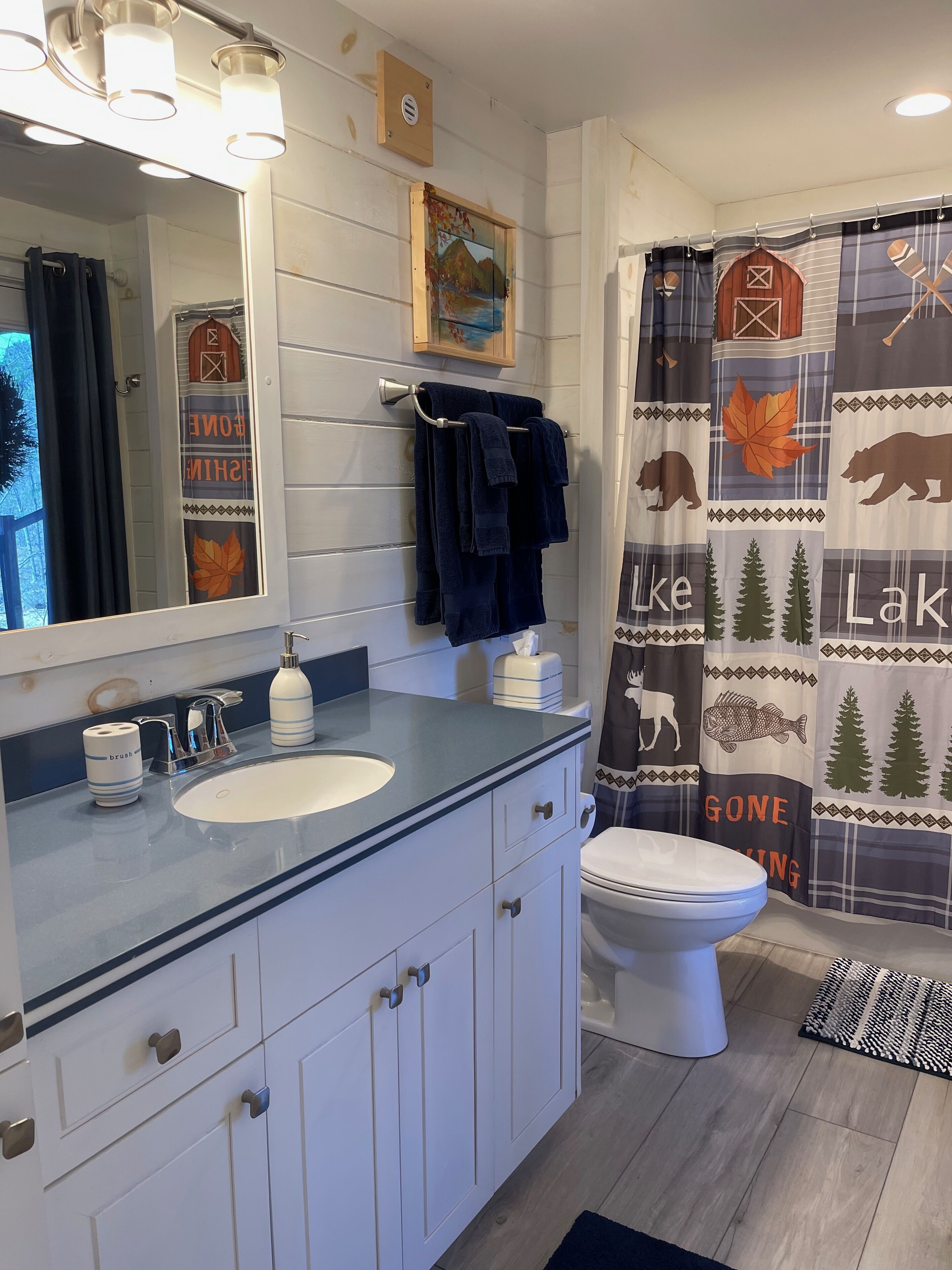 A bathroom with a white sink, a mirror, and a toilet. There is a towel rack with towels, a shower curtain with a nature theme, and a small shelf with decorative items.