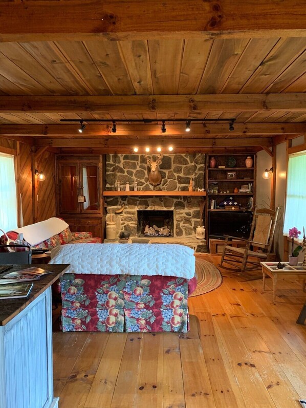 A cozy cabin interior with a stone fireplace, wooden beams, a bed with floral bedding, and a bookshelf.