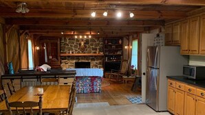 Een houten interieur met een eettafel, stoelen, een stenen openhaard en een keuken met een koelkast en magnetron.