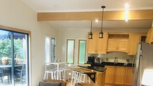 Uma sala de estar ampla com um sofá, poltronas e uma mesa de centro. Cozinha visível ao fundo, com armários de madeira e uma geladeira.