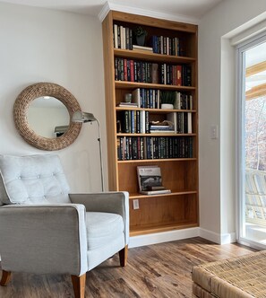A living room with a grey armchair, a wooden bookshelf filled with books, a round mirror, and a window with a view of trees.