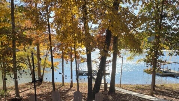 Um alpendre à beira do lago com cadeiras Adirondack e uma lareira, rodeado por árvores com folhagem outonal.
