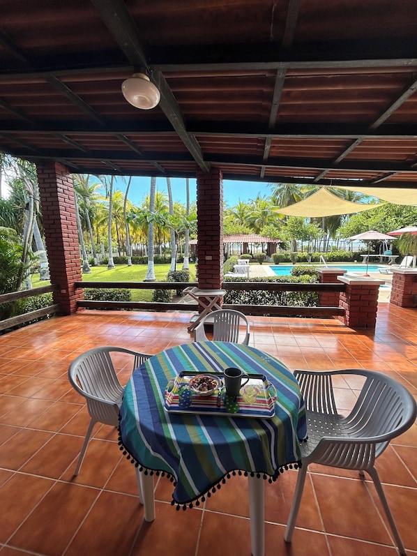 An outdoor patio with a table set for two, surrounded by chairs, overlooking a pool and palm trees.