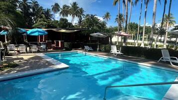 Piscina com espreguiçadeiras e guarda-sóis, cercada por palmeiras e um céu claro.