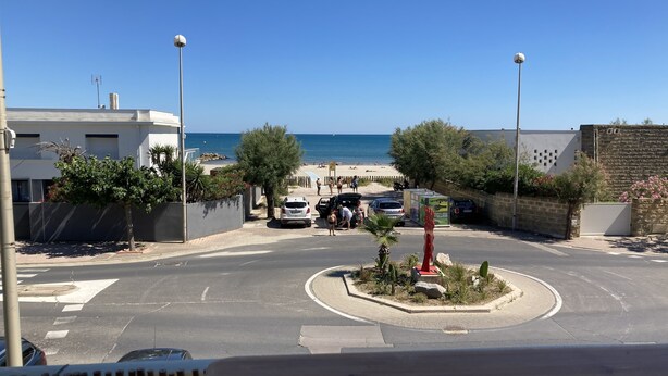 Straßenansicht mit Kreisverkehr, parkenden Autos, einem Strand im Hintergrund und blauem, wolkenlosem Himmel.