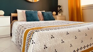 Ein Schlafzimmer mit einem Bett, einem runden Spiegel, einem Nachttisch, einer Topfpflanze und einem Fenster mit Vorhängen.